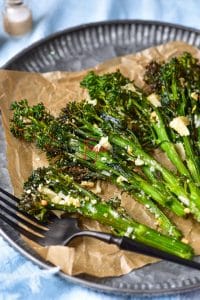 Air fryer broccolini are placed on a brown wax paper on a grey plate. The broccolini are crispy and have dark green florets and are covered with parmesan and garlic. A black fork is on the plate.