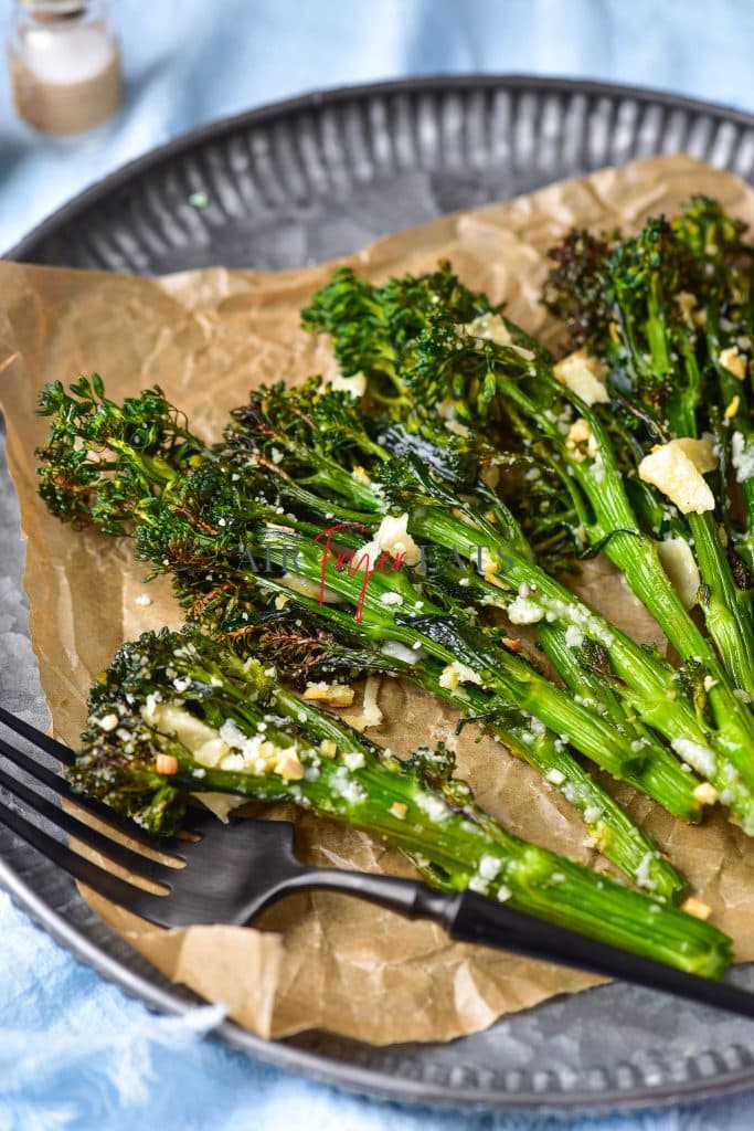 Air fryer broccolini are placed on a brown wax paper on a grey plate. The broccolini are crispy and have dark green florets and are covered with parmesan and garlic. A black fork is on the plate.