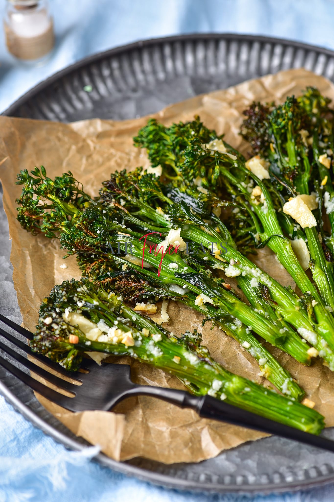 Air fryer broccolini are placed on a brown wax paper on a grey plate. The broccolini are crispy and have dark green florets and are covered with parmesan and garlic. A black fork is on the plate.