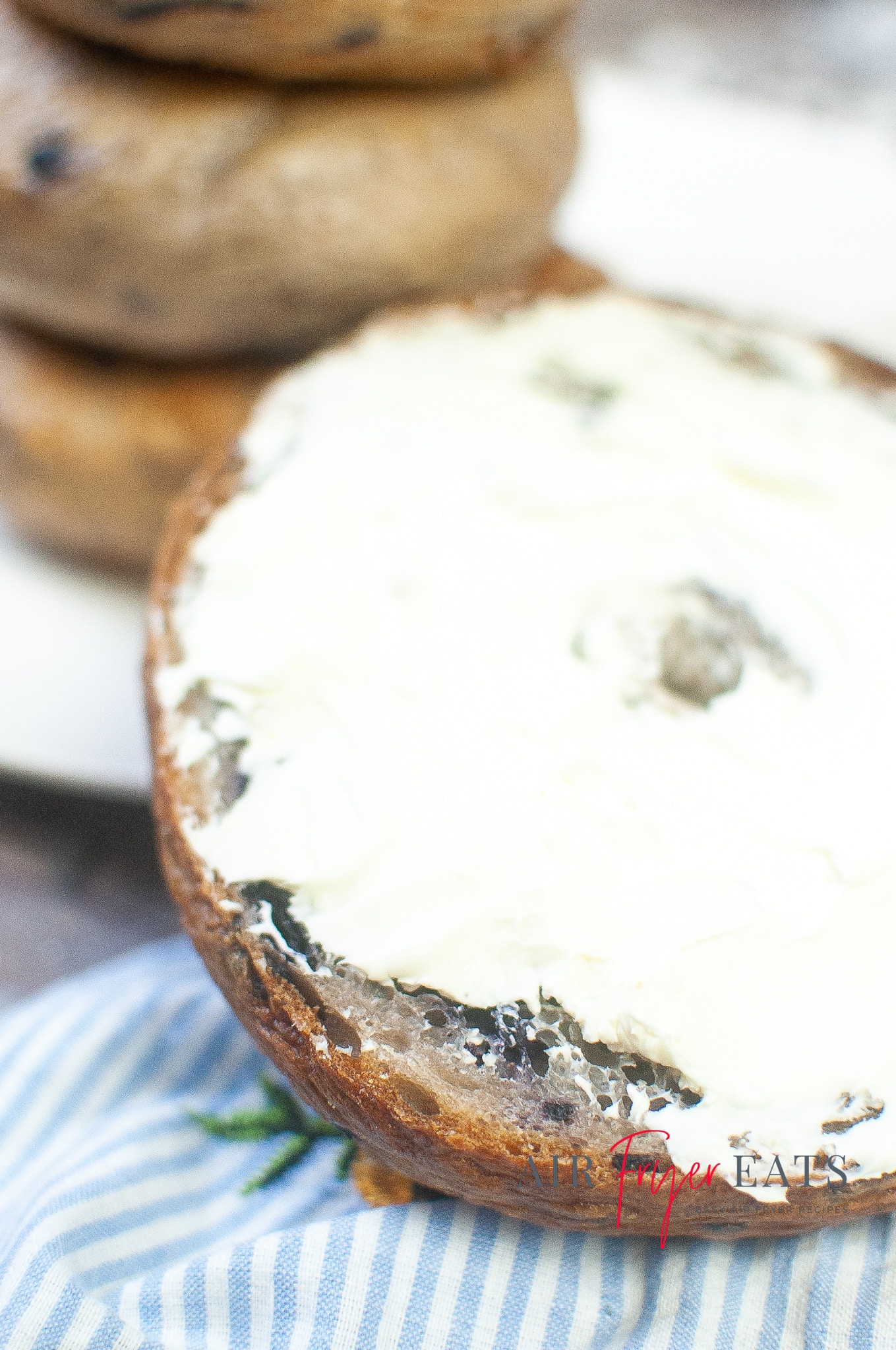 Close-up shot of an air fryer blueberry bagel that has been sliced in half and covered with white cream cheese. The bagel rests on a blue and white striped kitchen towel.