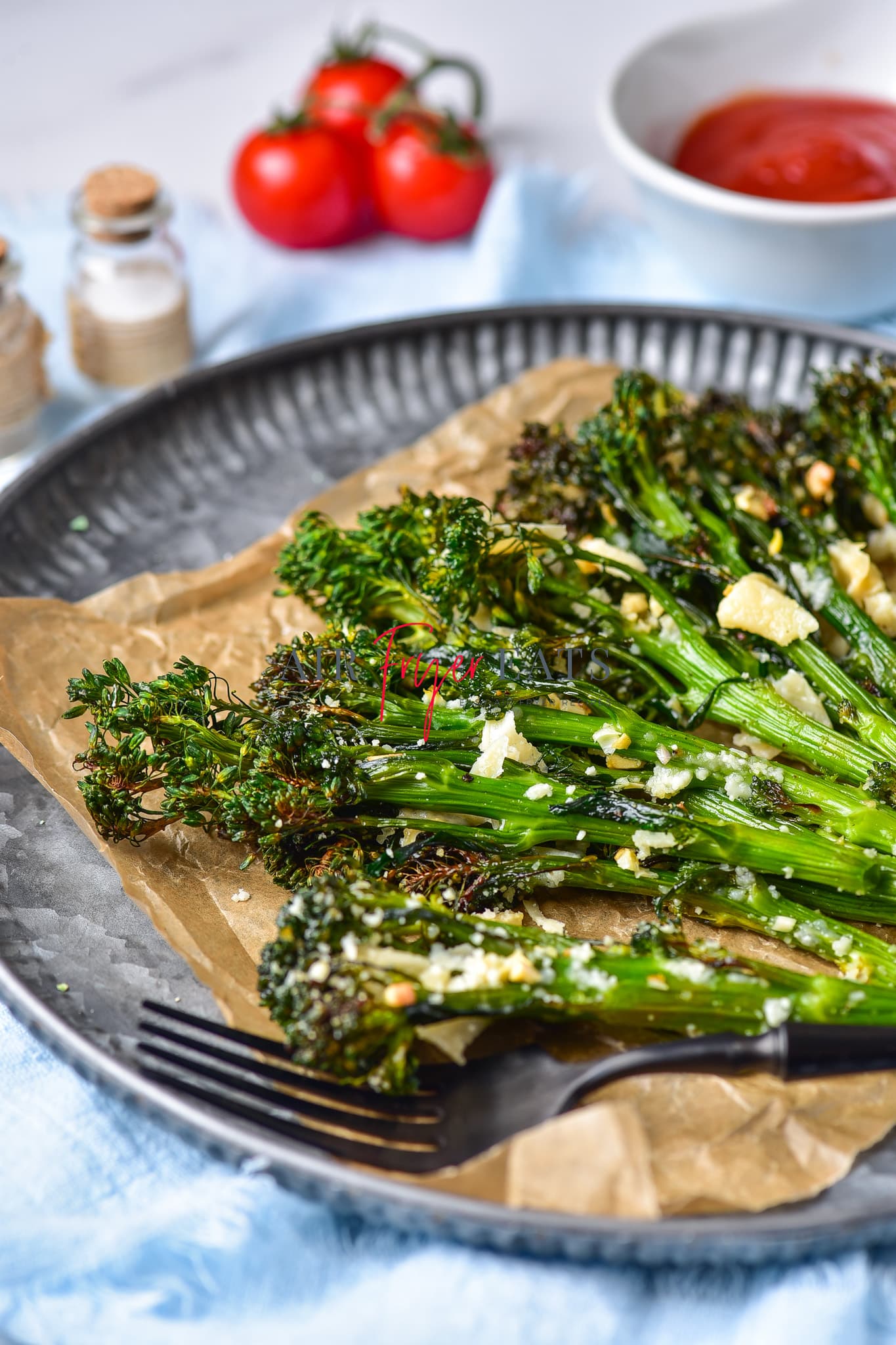 Broccolini in air fryer are placed on a brown wax paper on a grey plate. The broccolini are crispy and have dark green florets and are covered with parmesan and garlic. A black fork is on the plate.