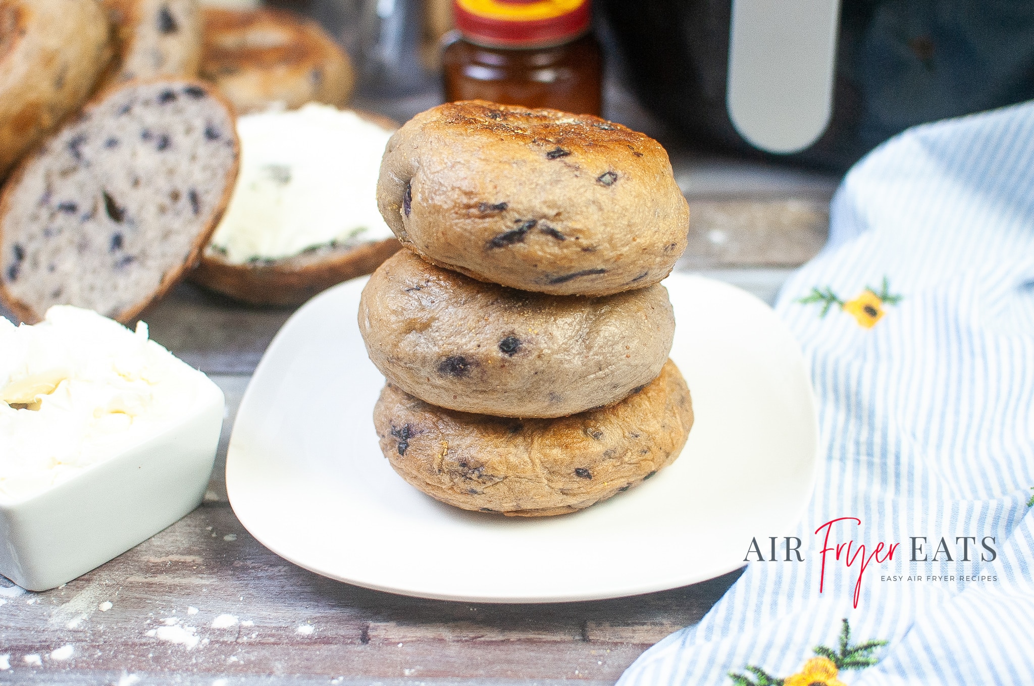 A stack of three perfectly cooked air fryer bagels are plated on a white plate. The top of the bagel is a rich brown. Nearby, there are sliced bagel halves, a black air fryer, and a blue and white striped kitchen towel.