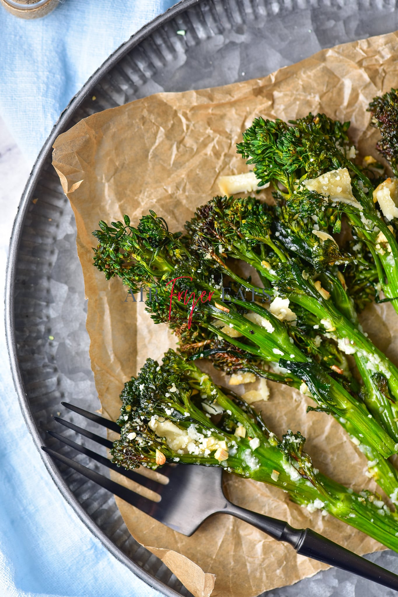 A close up shot of cooked broccolini in air fryer placed on a brown wax paper on a grey plate. The broccolini are crispy and have dark green florets and are covered with parmesan and garlic. A black fork is on the plate.