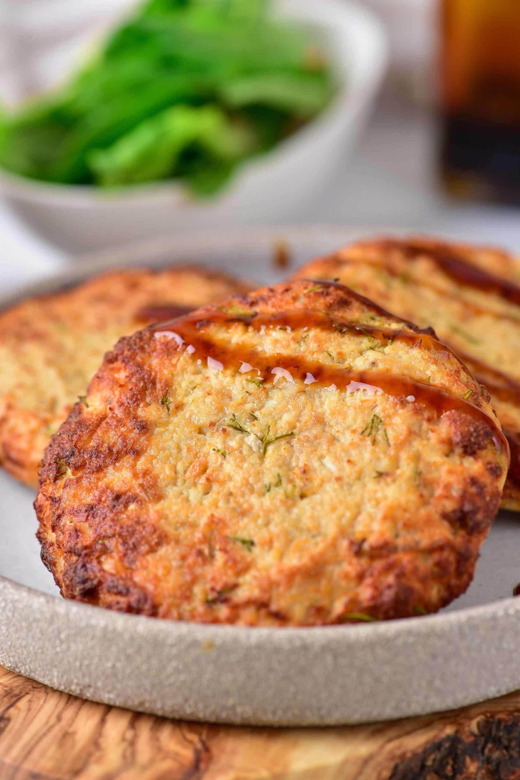Three golden-brown air fryer turkey patties are placed on a gray plate. They are covered with a little bit of barbecue sauce. In the background, there is fresh green lettuce.