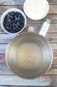 A large metal bowl is full of water with yeast. Nearby, there are two small bowls filled with frozen blueberries and flour.