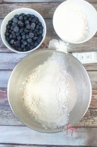A large metal bowl is full of water with yeast and flour, ready to be mixed. Nearby, there are two small bowls filled with frozen blueberries and flour.