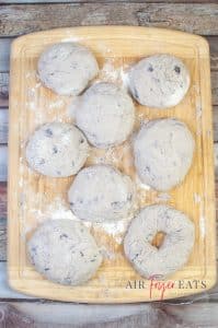 Bagel dough is formed into small balls on a flour-coated wooden board. There are seven balls of blueberry bagel dough and one one bagel shaped dough in the bottom corner of the wooden board.