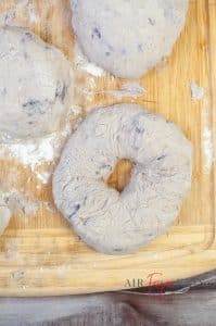 Bagel dough is formed into small balls on a flour-coated wooden board. A close-up image of one blueberry bagel is in the foreground, and a few balls of bagel dough are in the background.
