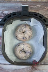 Two uncooked blueberry bagels are placed atop wax parchment paper inside an air fryer basket, ready to be cooked.