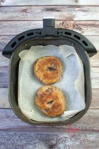 Two cooked air fryer blueberry bagels are sitting inside an air fryer basket on wax parchment paper.