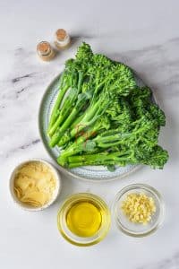 Freshly washed broccolini is plated next to a small bowl of parmesan flakes, olive oil, and minced garlic.