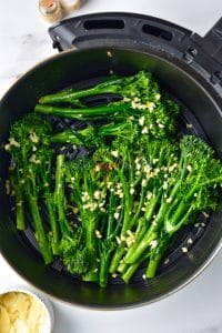 Air fryer broccolini is placed in a round air fryer basket. The green broccolini are covered in minced garlic.