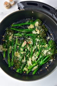 Cooked broccolini are covered in parmesan flakes and garlic peices in a round, black air fryer basket.