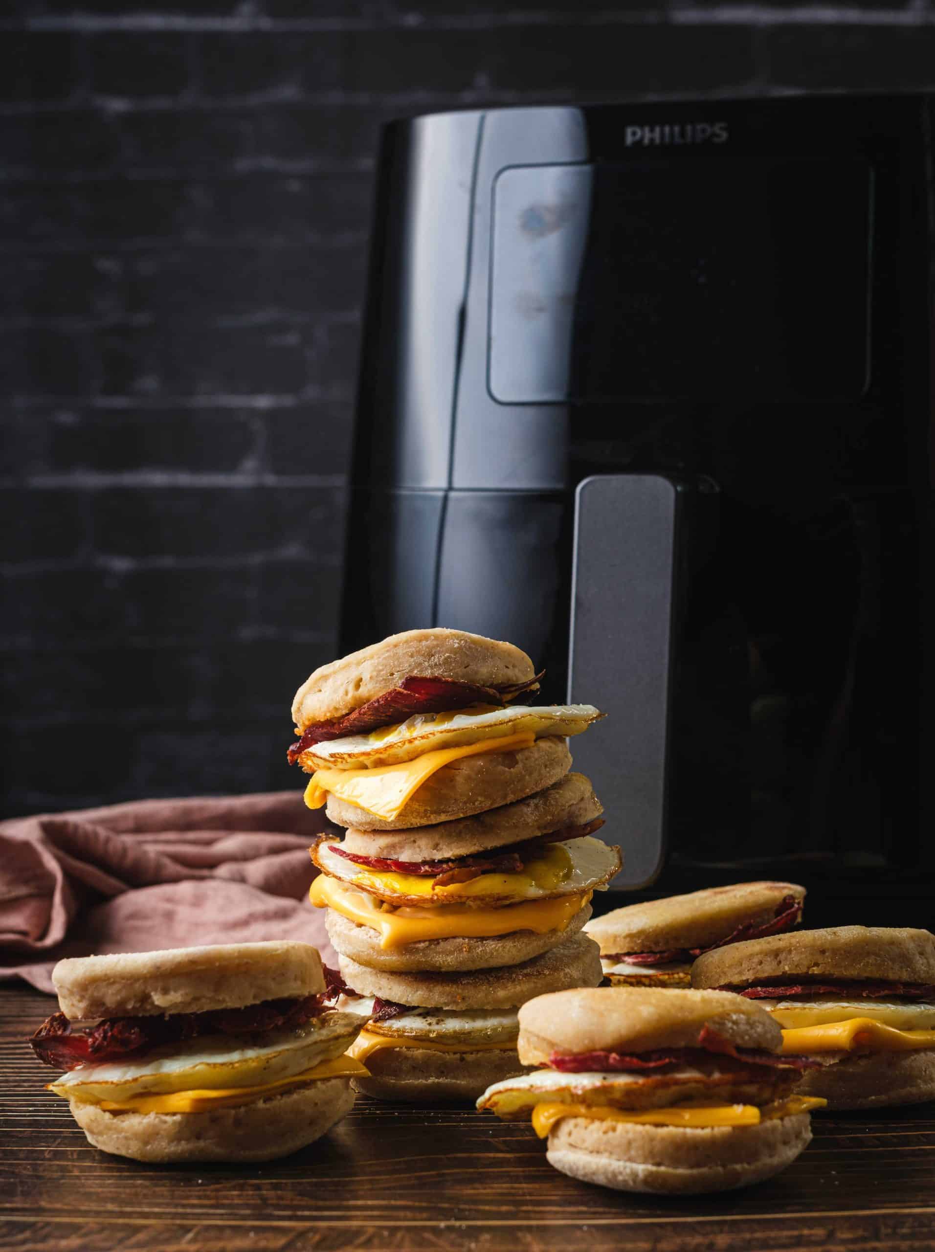 Air fryer Egg McMuffins are filled with Canadian bacon, a fried egg, and cheddar cheese. The breakfast sandwiches are stacked one on the other on a wooden kitchen board in front of a large, black Philips air fryer.