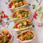 Shrimp tacos made in air fryer. There are five small tacos with flour tortillas, jalapenos, limes, cilantro, red onion, and air fryer shrimp. The tacos are colorful and beautiful and placed in a metal taco holder. One taco is laid on a wooden grey table.