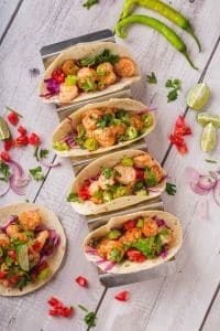 Shrimp tacos made in air fryer. There are five small tacos with flour tortillas, jalapenos, limes, cilantro, red onion, and air fryer shrimp. The tacos are colorful and beautiful and placed in a metal taco holder. One taco is laid on a wooden grey table.