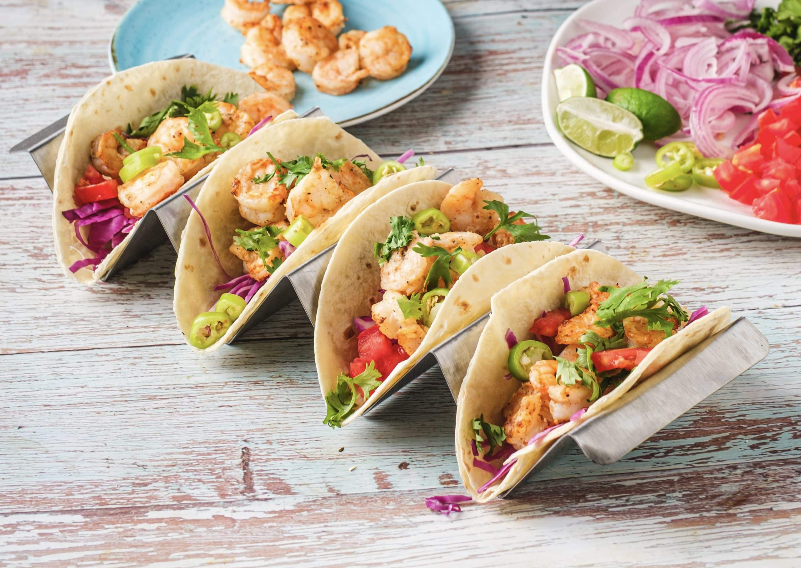 Four air fryer shrimp tacos are placed in a metal taco holder. The shrimp is orange and perfectly cooked. The colorful tacos are filled with purple cabbage, cilantro, tomatoes, and jalapenos. Behind the tacos, there is a plate of air fried shrimp and a plate of taco fillings with green limes.