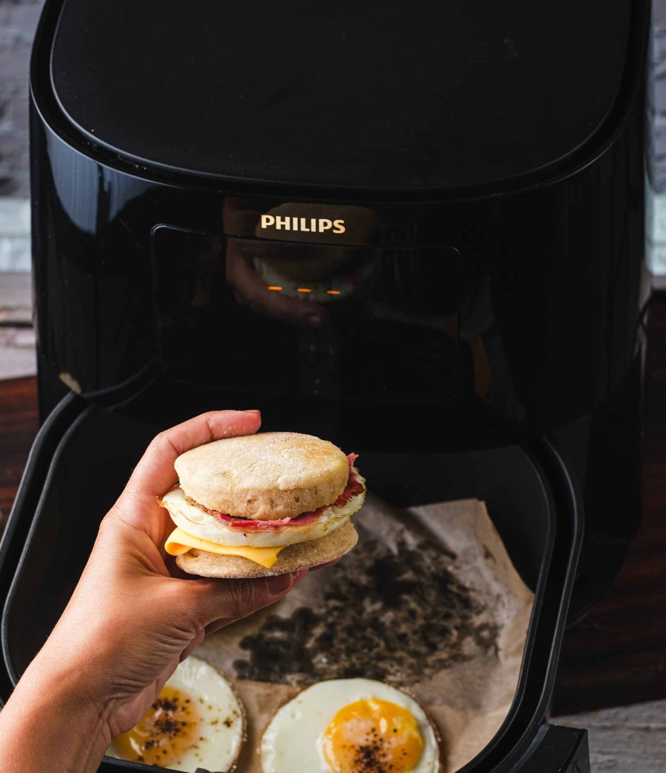 A home chef holds up an air fryer Egg McMuffin in front of an open air fryer basket that contains two fried eggs.