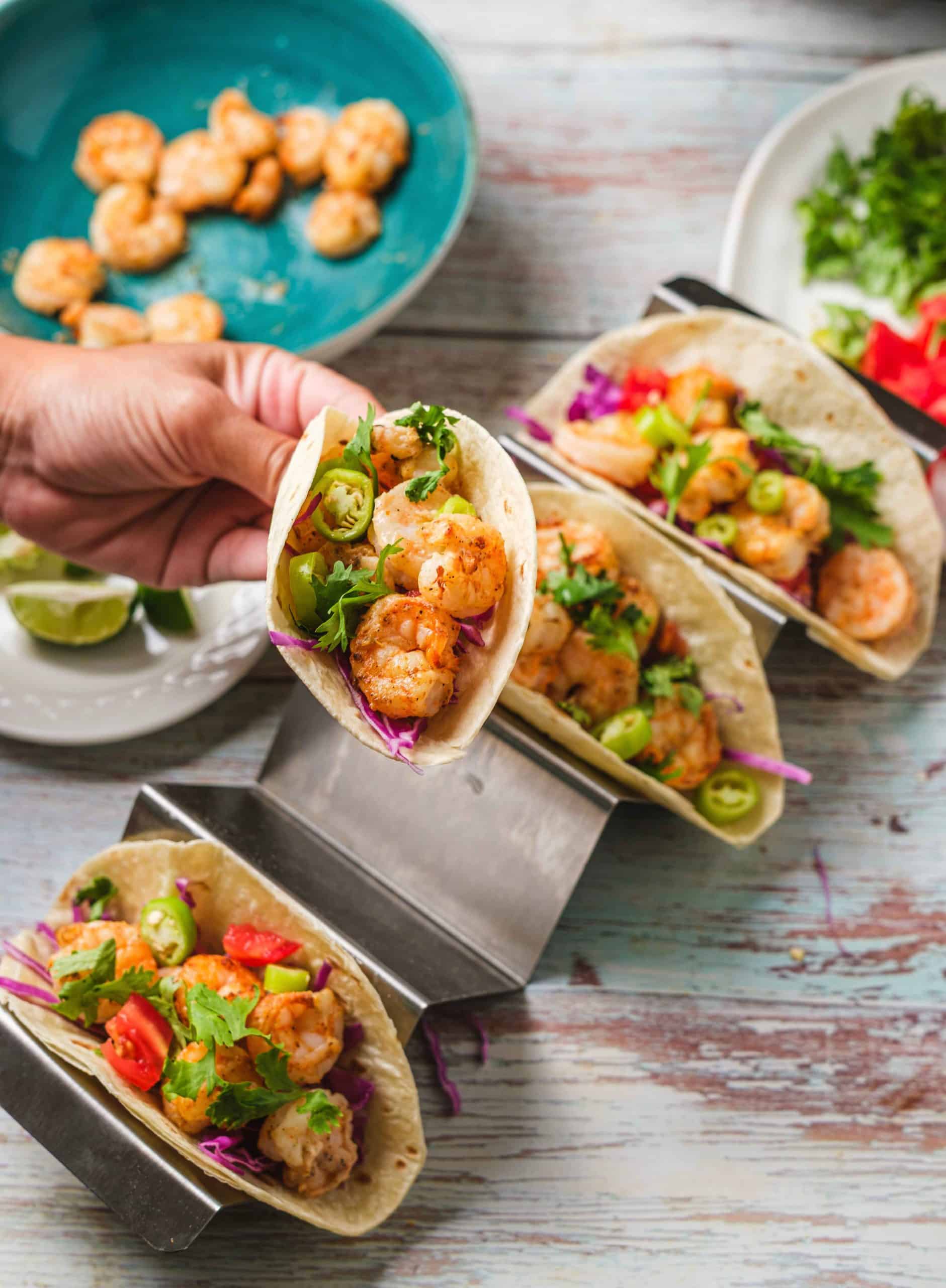 A chef holds an air fryer shrimp taco. The shrimp is orange and cooked perfectly. The tacos are filled with jalapenos, cilantro, tomatoes, cabbage, and onions. In the background there are three additional tacos, a plate of limes, a plate of shrimp, and a plate of taco fillings.