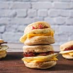 Four air fryer Egg McMuffins sit on a wooden cutting board. Each is fulled with Canadian bacon, a fried egg, and cheddar cheese.