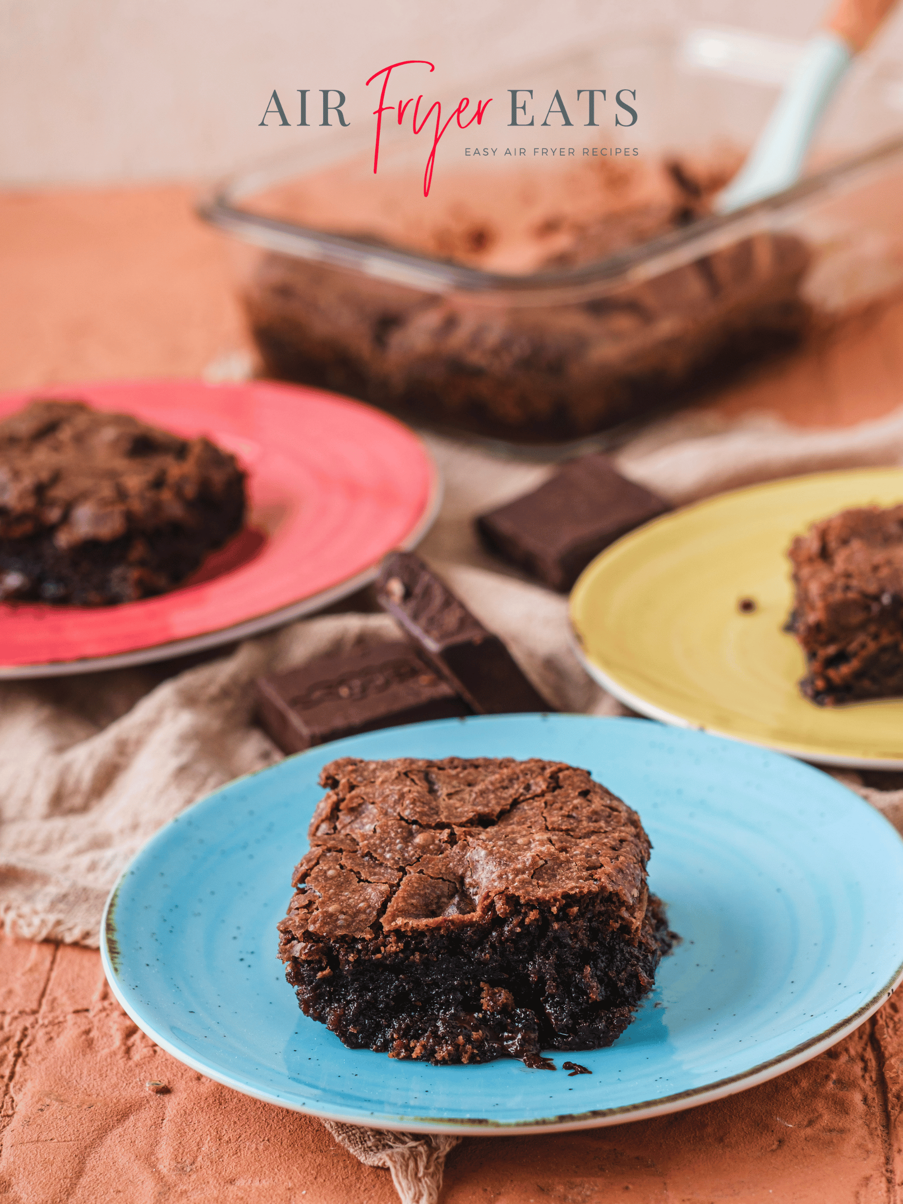 Three small, colorful plates have air fryer brownies placed on them. The brownies are gooey inside and have a crinkly top. In the background, there is a small glass container of brownies with a spatula for serving them.