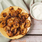 A plate full of air fryer tostones. The fried plantains are a dark yellow color with caramelized edges. They are next to a bowl of a white dipping sauce.