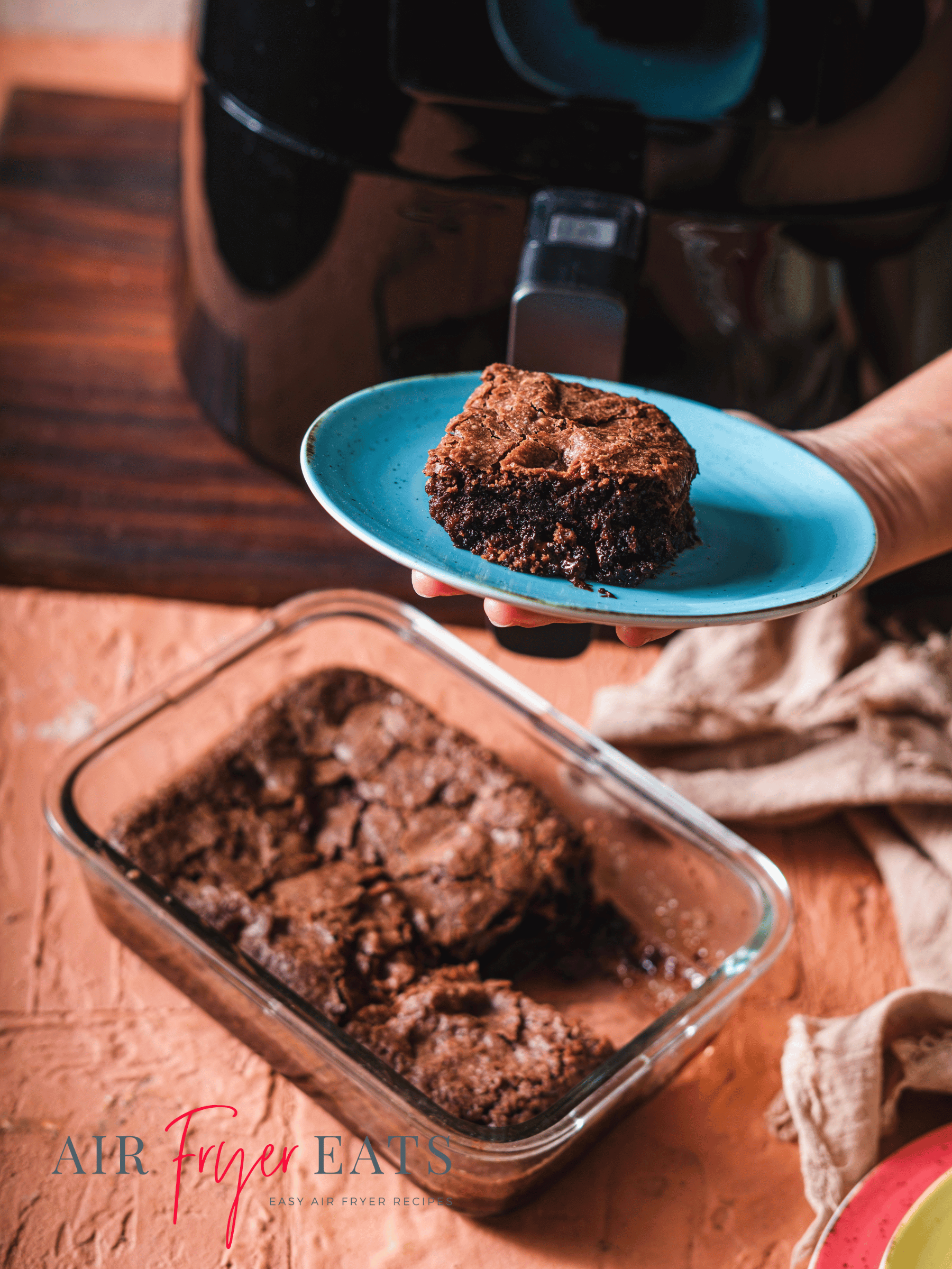 A home chef holds up a blue plate with a large air fryer brownie on it. Below, a glass baking container has the rest of the air fryer brownies. A large, black air fryer is in the background.