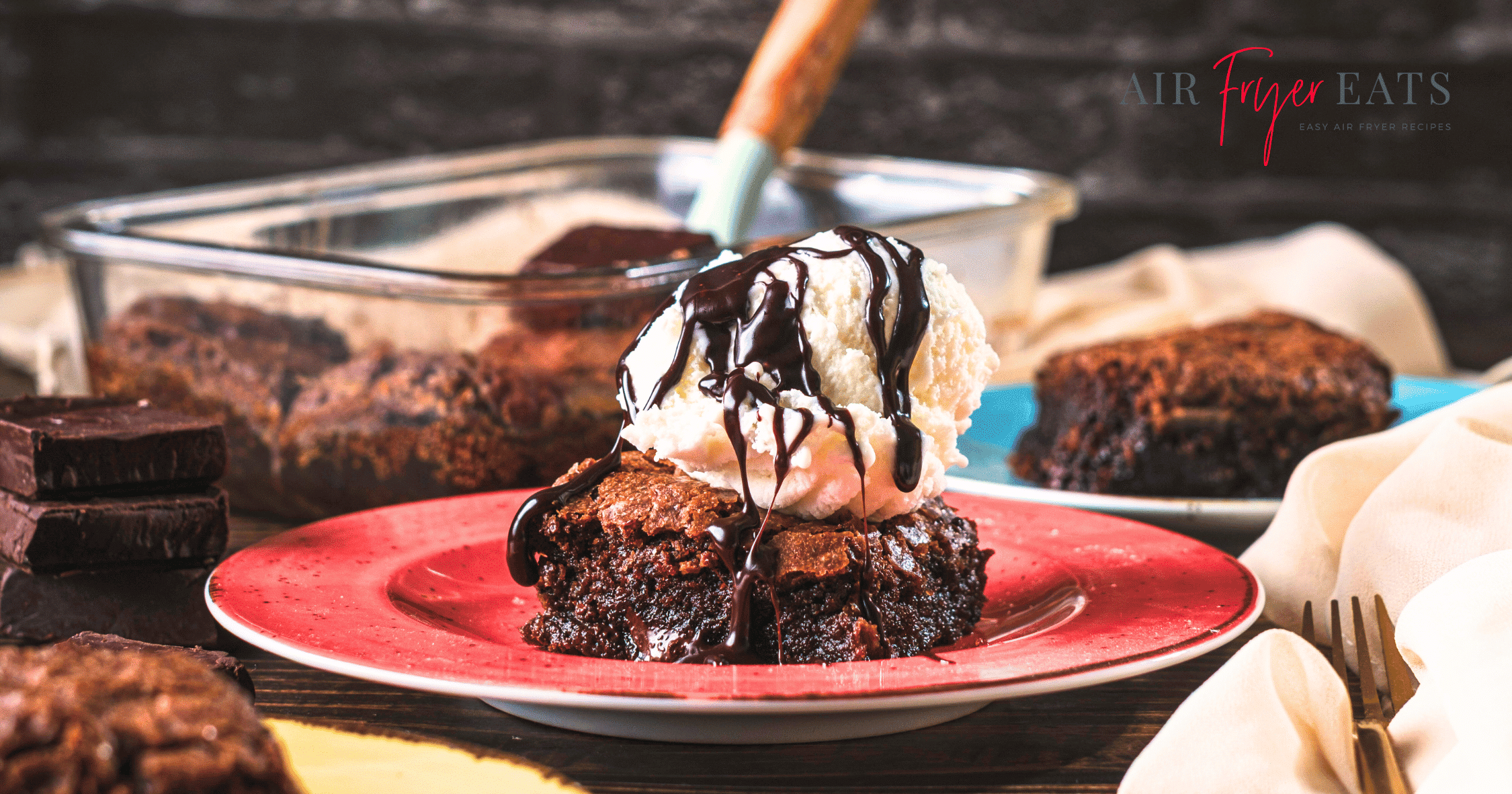 A delicious, gooey brownie with vanilla ice cream on top. Chocolate syrup drips down the ice cream onto the brownie. In the background, there is another plate with a brownie on it, a glass container full of air fryer brownies, and a chocolate pieces.