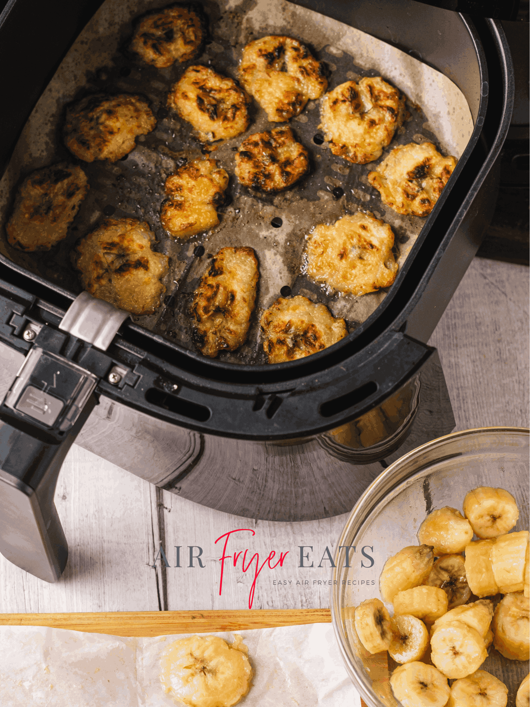 An air fryer is full of small fried plantains. The plantains are crispy and sweet. They fill the air fryer basket. Nearby, there is a bowl of cut plantains and a cutting board where the pieces are flattened.