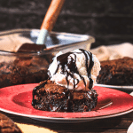 A gooey air fryer brownie is covered with a scoop of vanilla ice cream. Chocolate syrup drips down the ice cream onto the brownie. In the background, there is a glass baking container full of brownies made in the air fryer.