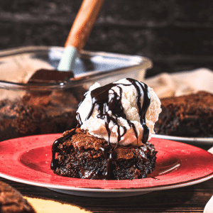 A gooey air fryer brownie is covered with a scoop of vanilla ice cream. Chocolate syrup drips down the ice cream onto the brownie. In the background, there is a glass baking container full of brownies made in the air fryer.