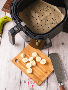 A green banana is peeled and chopped into small pieces on a wooden cutting board. There is a large air fryer basket in the image and a large knife.