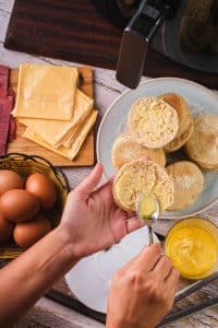 A home chef spreads butter on both sides of a sliced English muffin. In the background, the rest of the air fryer Egg McMuffin ingredients can be seen: cheddar cheese, Canadian bacon, and eggs, along with the air fryer.