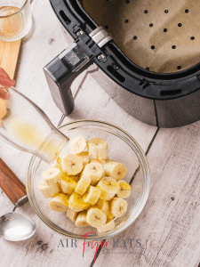 A large glass bowl of banana pieces is drizzled with olive oil. There is a large black air fryer nearby.
