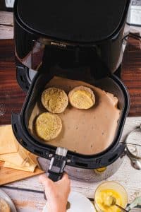 An air fryer basket contains three halves of English muffins. They are buttered and ready to be toasted in the air fryer.