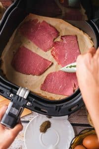 There are four slices of Canadian bacon in a rectangular air fryer basket. A chef is moving them with a kitchen tool.