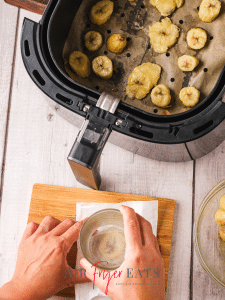 On a cutting board, a home chef uses a glass cup and parchment paper to flatten a fried plantain piece. A nearby air fryer basket is full of plantain pieces, some are flattened and some are not.
