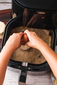 A home chef breaks an egg into an egg mold inside an air fryer basket to make air fryer breakfast sandwiches.