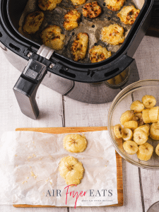 A few plantain pieces are placed on parchment paper on a cutting board, they have been flattened and are ready to be double-fried in the air fryer. A glass bowl of chopped bananas is nearby.