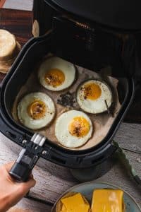 An air fryer basket is filled with four circular fried eggs. They have a bright yellow yolk and a white exterior and are sprinkled with black pepper. Nearby, there are English muffins and cheddar cheese to make an air fryer breakfast sandwich.