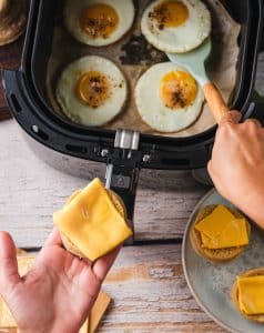 A home chef carefully removes fried eggs from an air fryer basket an prepares to place them on a cheese-covered half of an English muffin to make a breakfast sandwich.