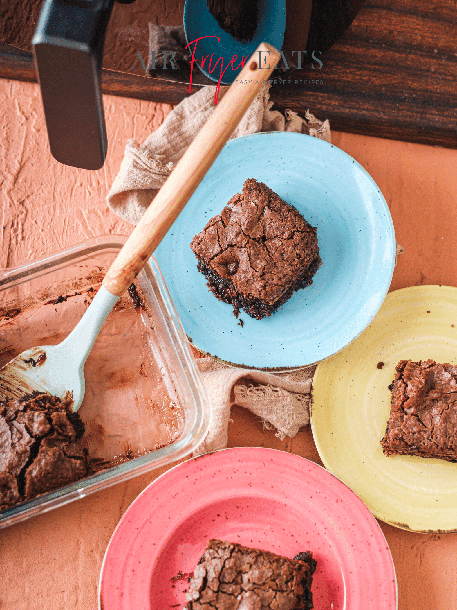 Three colorful plates with air fryer brownies on them lie next to a glass baking tray full of brownies. The brownies have a cracked top and gooey interior. There is an air fryer nearby.
