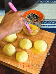 A home chef spreads egg wash atop bagel bite dough balls. There is a small plate of black and white sesame seeds nearby ready to be spread on the bagel bites as well.