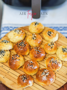 A round wooden board is covered with cooked air fryer bagel bites. They are golden brown and covered with white and black sesame seeds. There is a large black air fryer in the background.