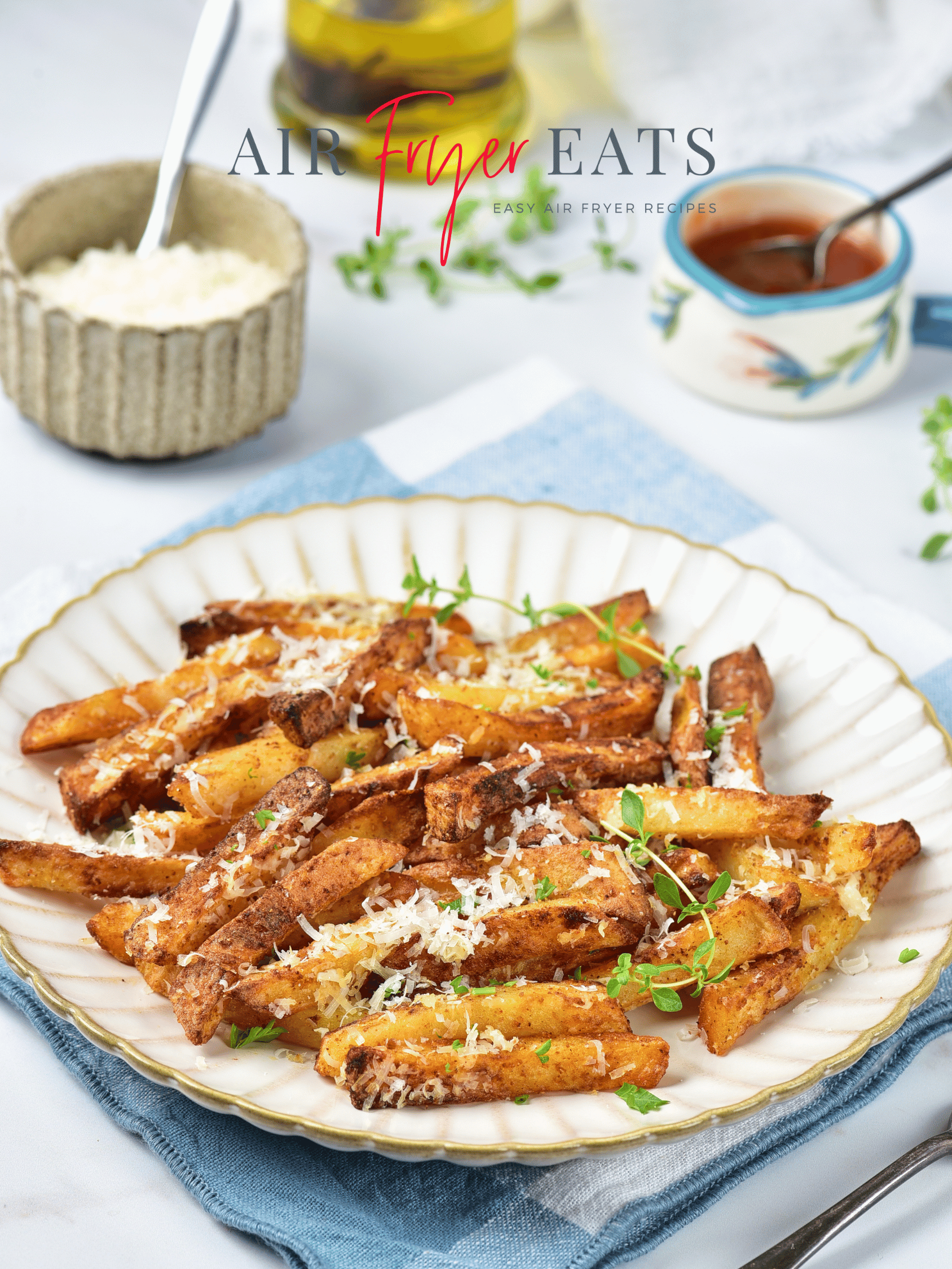 A white plate with gold edges is filled with crispy golden brown garlic parmesan fries made in the air fryer. The plate is set atop a blue and white kitchen towel. There is a small bowl with red sauce, a bowl with parmesan cheese, and a container of olive oil nearby.