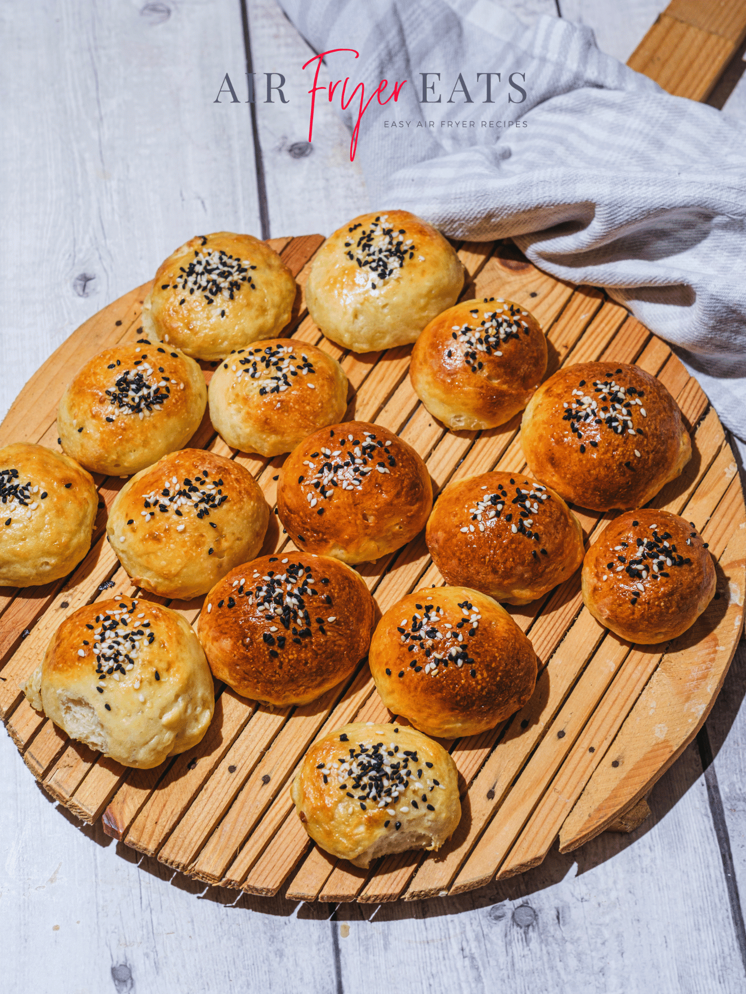 A round wooden board is covered with cooked air fryer bagel bites. They are golden brown and covered with white and black sesame seeds. The wooden board is set atop a grey wooden table and there is a decorative kitchen towel in the background.