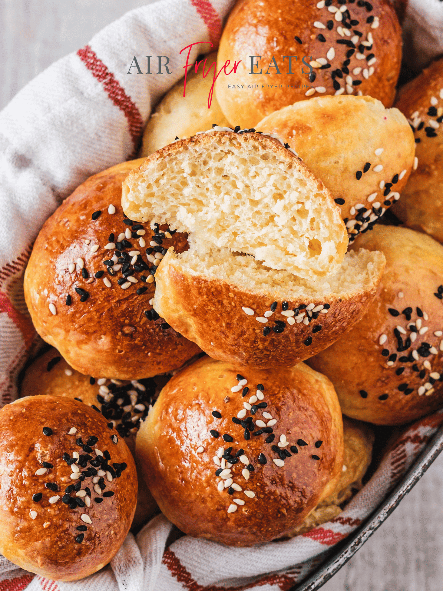 A tin basket of air fryer bagel bites. The bagel bites are golden brown and covered with black and white sesame seeds. There is one bagel bite sliced open, and the inside is fluffy and white. The bagel bites are atop a white and red striped kitchen towel.
