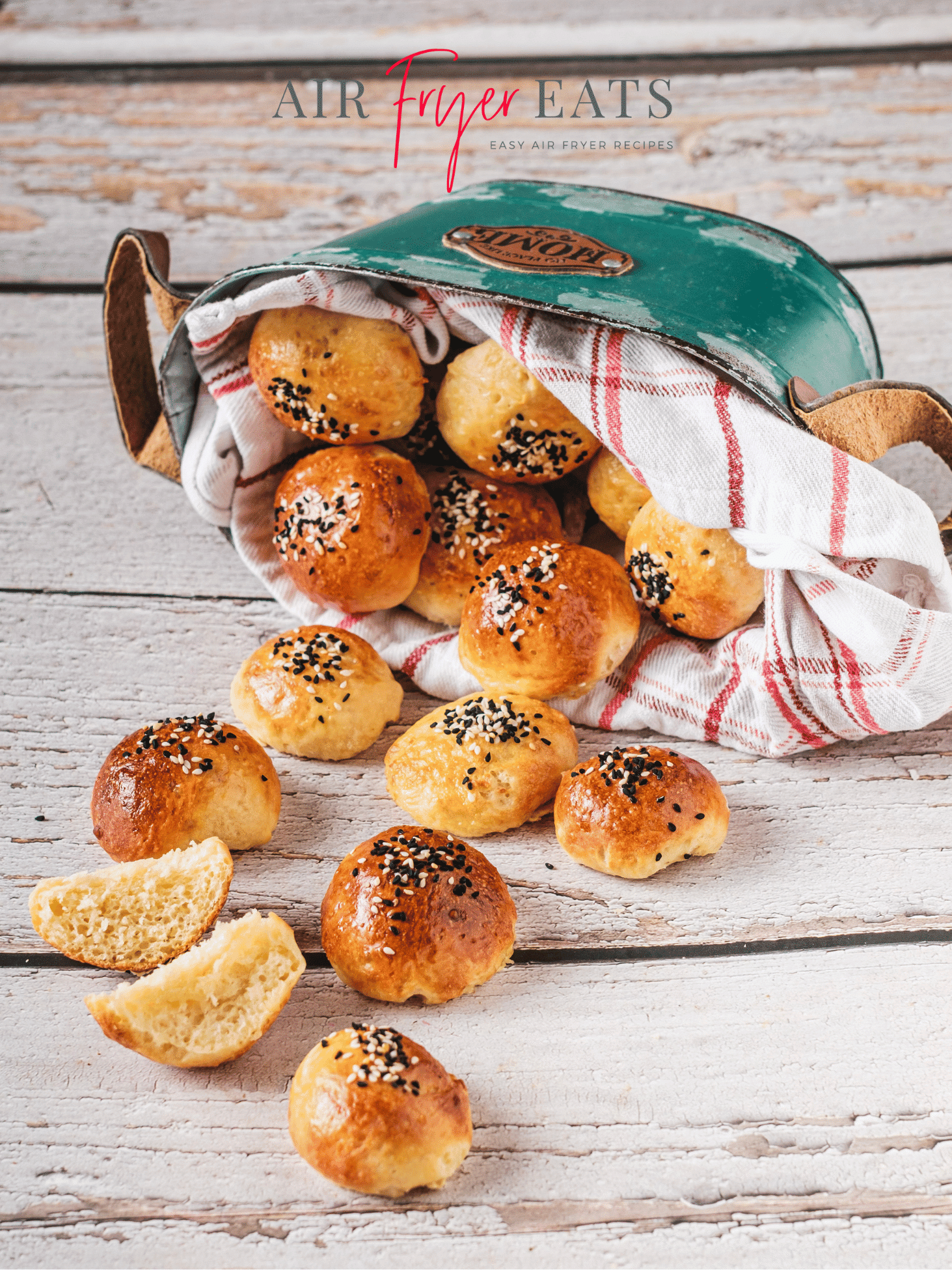A green tin basket of air fryer bagel bites is laying on a wooden countertop. The bagel bites are golden brown and covered with black and white sesame seeds. They fall out of the green tin and onto the wooden table. There is one bagel bite sliced open, and the inside is fluffy and white. The bagel bites are atop a white and red striped kitchen towel.