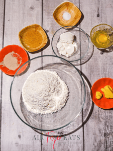 Bowls of air fryer bagel bites ingredients are placed on a grey wooden table. There is a glass bowl of flour, one of greek yogurt, two wooden bowls filled with honey and salt, and two orange bowls filled with yeast and butter. There is also a small glass bowl that has one egg.