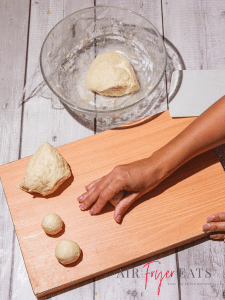 A home chef rolls dough into air fryer bagel bites. There are two small balls of dough already rolled and ready to be air fried. There is a ball of bagel bite dough in a glass bowl nearby and on the wooden cutting board.